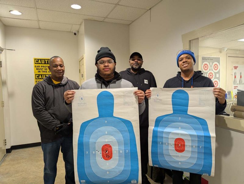 Four Down Range students at an indoor range, holding up the silhouette targets they shot during class.